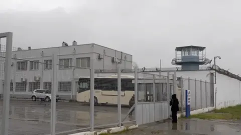 Rayhan Demytrie/BBC It is a grey day overshadowing a white-grey prison, which is a two storey, square block with rows of barred windows, surrounded by wire fencing with barbed wire. A coach and a car are waiting just inside the gates and a figure wearing black can be seen standing just outside them. It's raining.