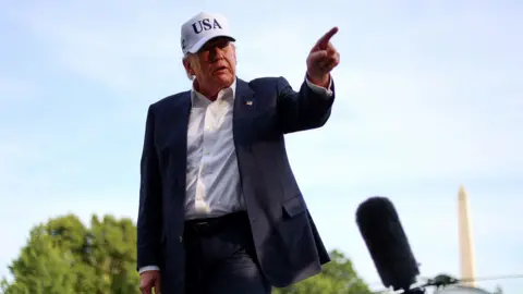 Alex WROBLEWSKI / AFP via Getty Images Trump wears a white USA cap and a dark suit, while pointing at something off-camera