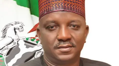 Nigeria's power ministry Saleh Mamman poses looking directly into the camera. He wears a grey top, a multi-coloured cap, and is pictured in front of a flag.