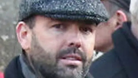 BBC Man looks at camera, he has dark stubble and is wearing a flat cap