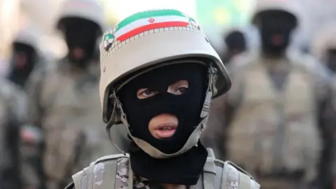 Majid Asgaripour/WANA/Reuters A young person wearing a balaclava, fatigues and a helmet sporting an Iranian flag looks off camera, with blurred military figures in the background.