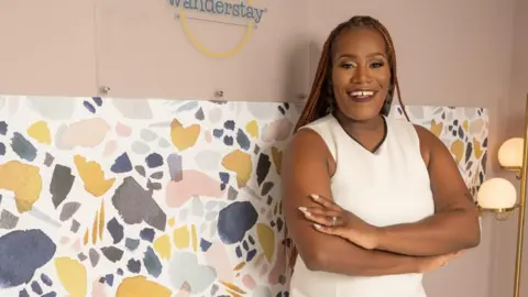 CJ Washington Deidre Mathis smiling, wearing a white dress leaning against a wall with printed paper and a pink backdrop saying Wanderstay