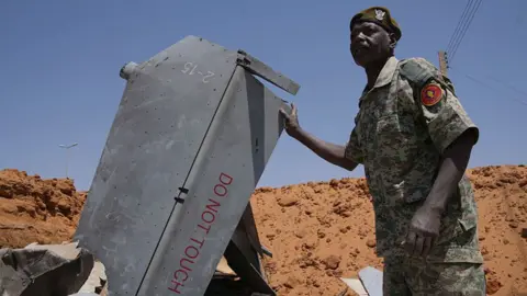 Los Angeles Times via Getty Images A man in a military uniform stands in a crater, surrounded by sand. His right hand rests on the fin of a downed drone which says 