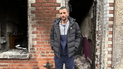 BBC A man with brown hair and a black puffer coat stands outside his home