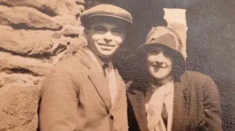 Le Breton family archive (Carolyn Horn) John and Phyllis Le Breton pictured smiling in an old sepia photograph. He is wearing a flat cap and suit and she is wearing a hat, coat and scarf. They appear to be standing in front of a stone monument.