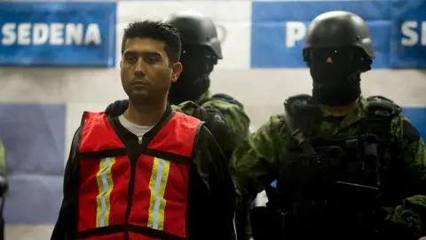 YURI CORTEZ/AFP via Getty Images In this file photo from 2012, Mexican soldiers are seen escorting Valencia Salazar after his arrest that year. He is wearing a high-vis vest over a dark hoodie and a white T-shirt. The two soldiers behind him are wearing helmets, dark glasses, and balaclavas. They are armed and are wearing bullet-proof vests. 