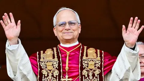 AFP via Getty Images Pope Leo XIV, Robert Prevost wearing ornate ceremonial robes with gold embroidery stands with both arms raised. He is smiling and wearing glasses.