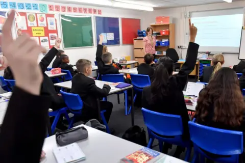 Anthony Devlin/Getty Images Several pupils seated at desks in the foreground raise hands, with exercise books and pencil cases visible on the tables. A teacher stands at the front beside a projected screen showing a lesson slide. Blue plastic chairs and light-grey desks are arranged in rows. Colourful display boards and number charts run along the wall above a green board. Shelving units hold plastic storage trays and stacked folders near the teacher’s desk.