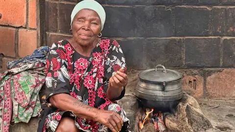 Adaobi Tricia Nwaubani Lami Ezekiel in front of the home built for her by the government in the late 80s. She appears to be blind in one eye and is wearing a black and red floral dress, She is sitting on the ground next to a saucepan on a wood fire.