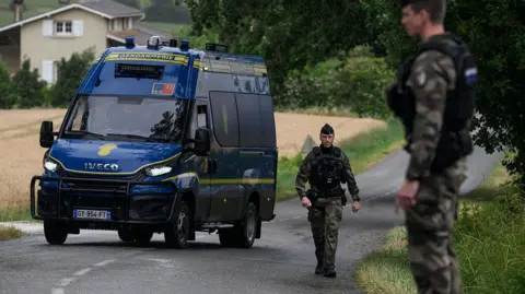 Getty Images French police officers stand near their van on a road outside a french country village during the search for the missing girl. The road cuts through fields and some buildings can be seen in the background.
