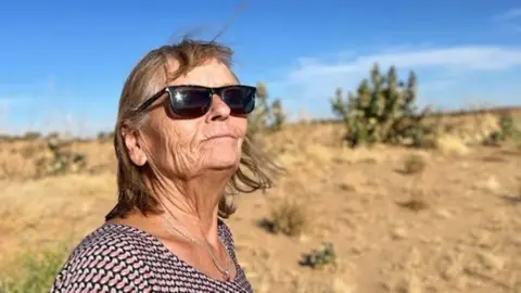 Christoph Greztmacher Eva Greztmacher looks up in sunglasses. A blue sky and desert scrubland can be seen behind her. She is wearing a necklace with a ring as a pendant.