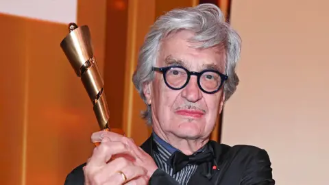 Getty Images Wim Wenders with grey hair and black glasses holding up an award