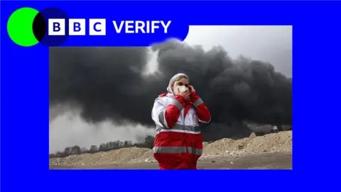 BBC An emergency worker in Tehran shields her face as black smoke rises behind her. She is wearing a red and white jacket and a face mask and standing in a sandy location. The image is imposed over the BBC Verify colours and branding. 
