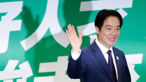 Reuters Taiwanese President Lai Ching-te stands to the right of the picture waving and smiling at his party HQ in Taipei. Behind him is a green backdrop with Chinese lettering.