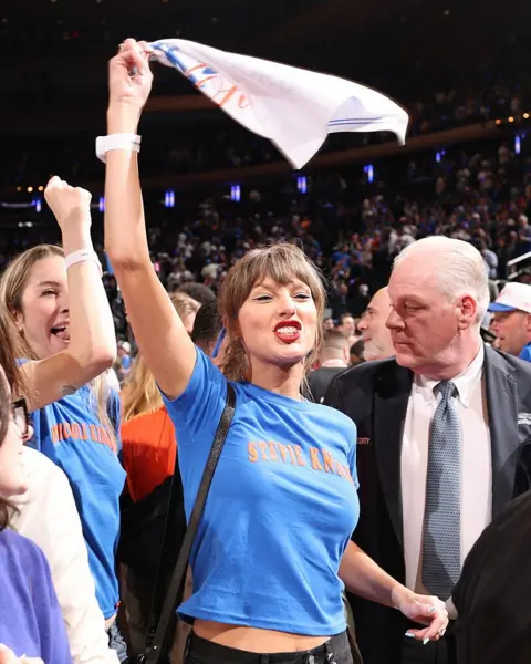 Getty Images Taylor Swift smiles and cheers as she waves a white towel in the air in excitement. In the background are other Knicks fans.