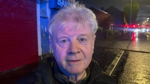 A man in close up standing outside on a rain-slicked street at night. Street lights, houses and trees can be seen in the background as well as shuttered businesses. The man is in his 50s or 60s, has short grey hair and is wearing a coat.