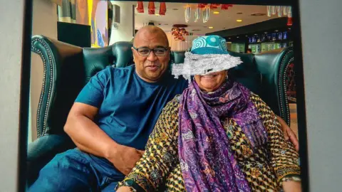 Netflix Pastry chief Graham Hornigold in a navy blue t-shirt sits with his mother Dionne Marie Hanna, wearing a baju kurung, purple scarf, teal hat, on a couch in a restaurant