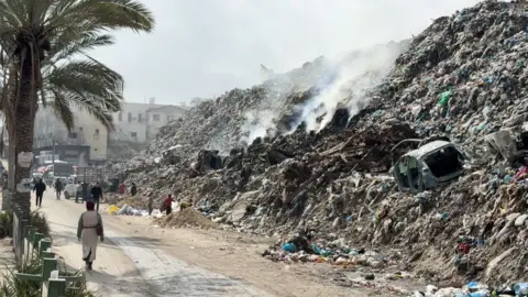 A mountain of debris steams in the heat on a street in Gaza. It's as high as the buildings and people walk by. Since the start of the war, most of Gaza's infrastructure has been destroyed.
