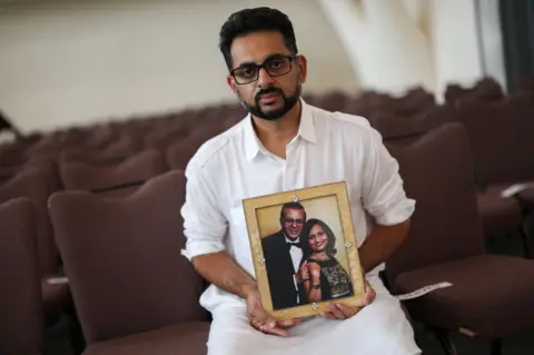 Reuters / Hannah Mackay A person in a white shirt sits among rows of empty auditorium seats, holding a framed photo of two people dressed in formal clothes. The background is softly out of focus, drawing attention to the person and the picture.