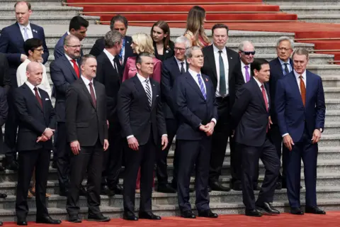 Getty Images Members of the U.S. delegation including (front L-R) White House Deputy Chief of Staff Stephen Miller, Trade Representative Jamieson Greer, Secretary of War Pete Hegseth, Secretary of the Treasury Scott Bessent, Secretary of State Marco Rubio, Ambassador to China David Perdue, and CEOs from various industries including Apple CEO Tim Cook, Tesla CEO Elon Musk and Nvidia CEO Jensen Huang, stand prior to a welcome ceremony for U.S. President Donald Trump at the Great Hall of the People on May 14, 2026 in Beijing, China.