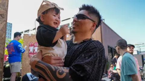 BBC/ Hosu Lee A man with a tattoo on his left arm is carrying a smiling toddler. 