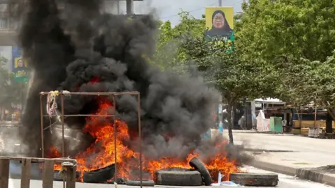 Reuters Burning tyres on a main road in Dar es Salaam with dark smoke rising from the flames. In the background a poster can be seen of President Samia Suluhu Hassan.