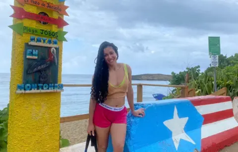 Supplied Debbie Perez standing beside a concrete barrier painted with the Puerto Rican flag next to a beach