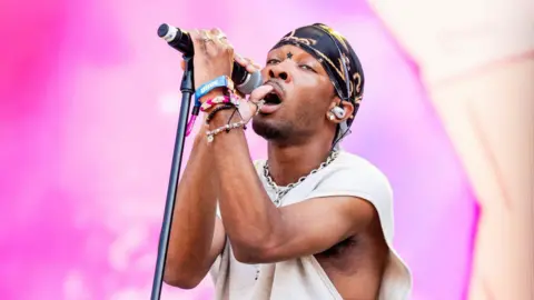 Getty Images D4vd holds a microphone stand close to him while performing onstage during a music festival in 2024. He has a black and gold headscarf on and a cream top.