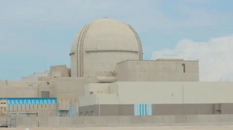 AFP A large sand-coloured building in the UAE called the Barakah Nuclear Power Plant. In the centre is a large dome while rectangular shaped sections of the building lie to the left, right, and in front of the dome. The sky behind is blue with a few white clouds.
