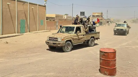 AFP via Getty Images FLA fighters sitting in a pickup truck on a sandy road, with another in the distance behind them. to the left are buildings lining the road. In the front right, there is an orange metal barrel
