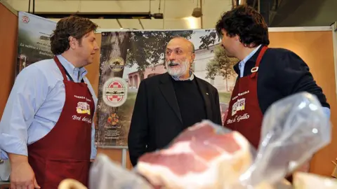Getty Images Petrini is in the centre of the image in a black suit and jumper, with two men in burgundy coloured aprons to each side of him. An unwrapped ham can be seen in the foreground of the picture out of focus.