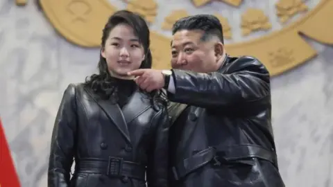 EPA With her long hair tied back, Kim Ju Ae, stands next to a pointing Kim Jong Un, wearing matching black leather belted jackets.