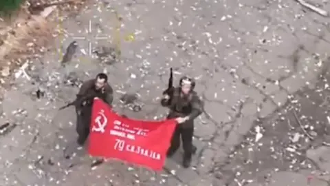 Russian defence ministry Two men hold a red flag with a hammer and sickle showing they are part of a Russian unit in a video posted by Russia's defence ministry