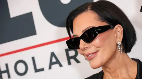 Variety via Getty Images Kris Jenner, wearing a pair of thick black sunglasses and dangling earrings, smiles for photographers at a red carpet event.