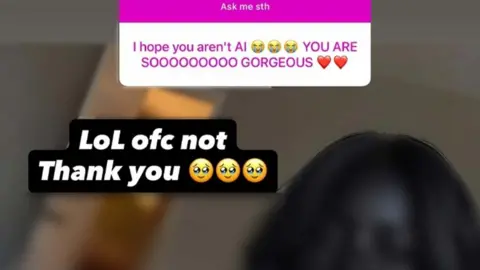 Instagram Screen shot taken from a Q&A on the Instagram account, which the BBC is not naming, that stole Riya Ulan's content. A comment reads: "I HOPE YOU AREN'T AI [three crying emojis] YOU ARE SOOOOOOOOO GORGEOUS [two red heart emojis]". A response is displaced saying: "LoL ofc not Thank you [three face emojis]. The head of a woman with black skin and hair is partially visible. The face and the backdrop have been blurred.