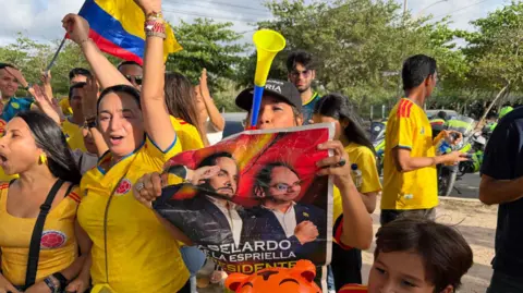 A crowd wearing yellow shirts with one person holding up a poster of Abelardo de la Espriella, celebrating on the streets with a Colombian flag in the background.