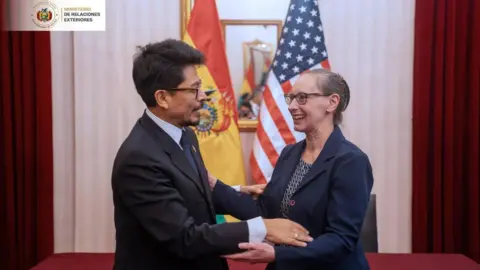 Cancillería Bolivia A man in a suit and glasses, with short dark hair, holds the arms of a woman wearing a shirt and suit jacket and glasses, in front of the American and Bolivian flags.