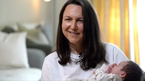 Ana is holding her baby, she is wearing a white top and has long brown hair.