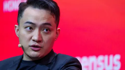 Getty Images Justin Sun in black blazer and black T-shirt, with small beige microphone and ear piece in front of red background