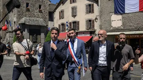 AFP via Getty Images Gabriel Attal (C) walks in streets of the village of Mur-de-Barrez during a visit in the Aveyron department