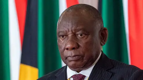 Bloomberg via Getty Images A head and shoulders image of Cyril Ramaphosa wearing a white shirt and red tie, with a pin-striped jacket. Behind him can be seen South African flags