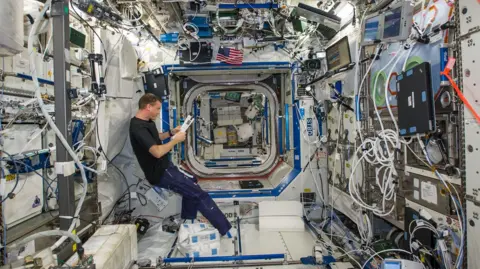 NASA Wiseman floats weightless inside a busy space station module, surrounded by equipment, laptops and tangled cables on every surface. He reads from a tablet while cargo bags, experiments and storage lockers line the walls and ceiling. The narrow, tunnel-like lab stretches away into connecting modules, lit by bright white panels. Every available space is packed with instruments, giving the high-tech interior a dense, cluttered feel ideal for microgravity research.
