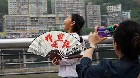 Visitors standing with their mouths open so it looks like they are "eating" the train. A woman doing the pose is also wielding an open white, red and black fan.