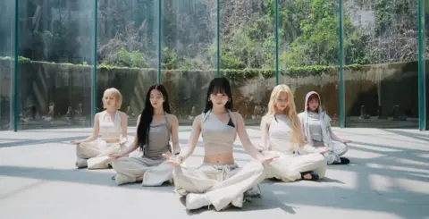 Source Music A still image from the performance video for Le Sserafim's song Boompala shows the band meditating in a plain room, whose windows look out onto a lush, mountainous backdrop