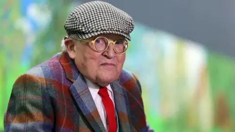 Getty Images David Hockney is seen in old age. He is wearing a check suit and a check cap and yellow round glasses. He is looking into camera.