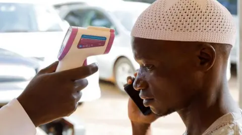 Reuters A man dressed in white is seen on the right of the image on his mobile phone as his temperature is taken with a contact-free temperature gun. A number of cars are out of focus in the background of the picture.