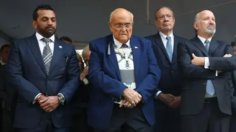 Getty Images Giuliani in center with blue suit jacket over white medical brace, crosses his hands and looks down. He is flanked by Kash Patel and Howard Lutnick