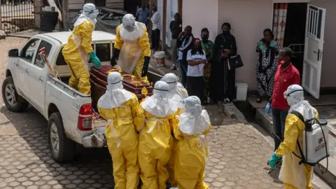 EPA Health workers wearing full personal protective equipment (PPE) lift a coffin of a victim on to the back of a pick-up truck in Bunia. Some people with masks watch them outside a hospital.