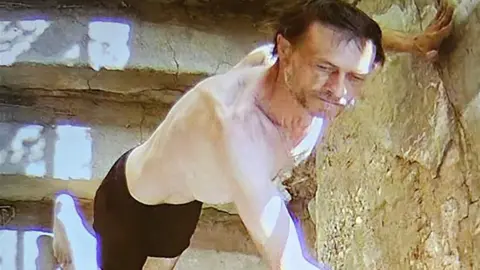 Reuters Pale-skinned man on light stone steps with facial hair stubble, unkempt hair, black shorts, presses his hands against nearby stone wall