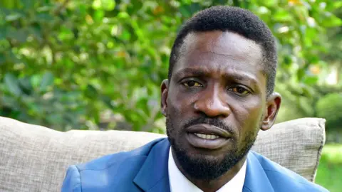 Reuters A close-up shot of Bobi Wine looking at the camera while sitting down. He is wearing a blue suit and has short hair and a beard.